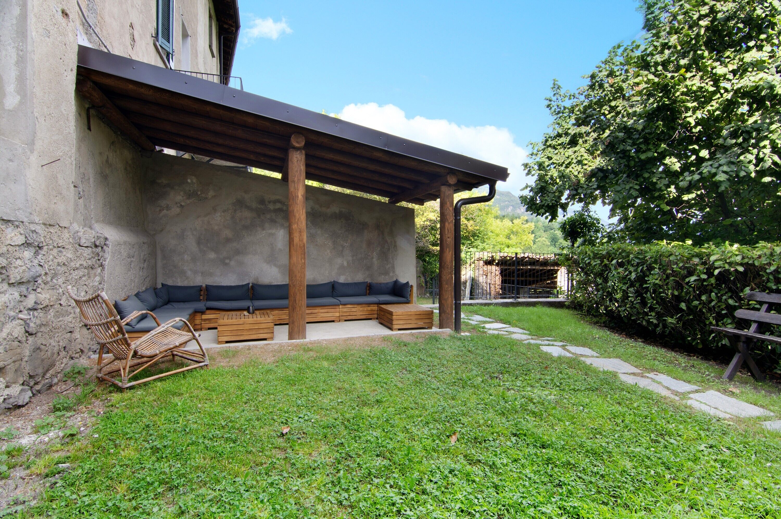 Un'area salotto all'aperto coperta, con una panchina e una sedia, un muro in pietra e un prato.