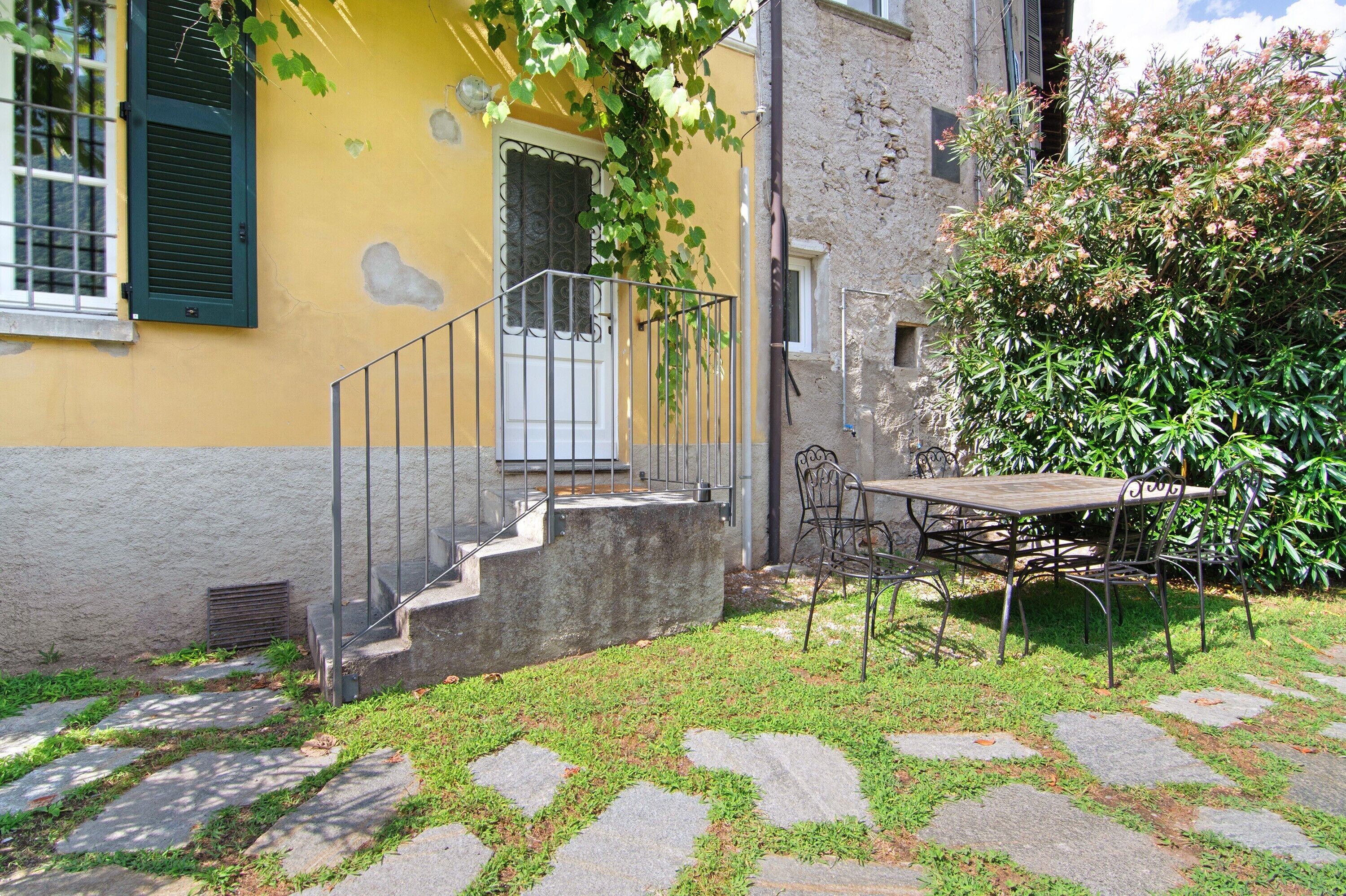 A yellow building with a staircase leading to a door, a table with chairs outside, and a garden area.