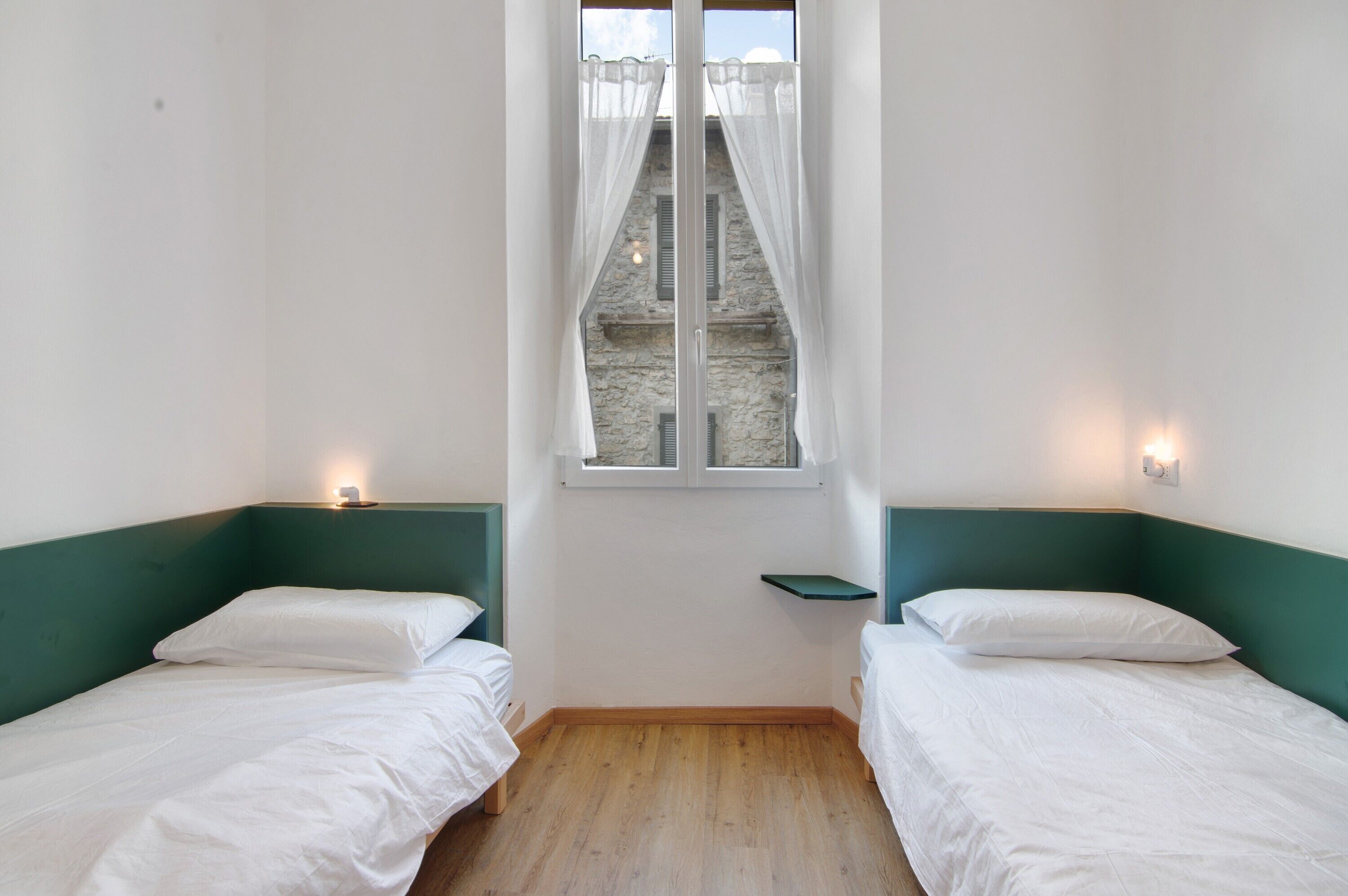 Une chambre avec deux lits simples, une fenêtre avec des rideaux légers, une petite étagère et une vue sur un bâtiment en pierre.