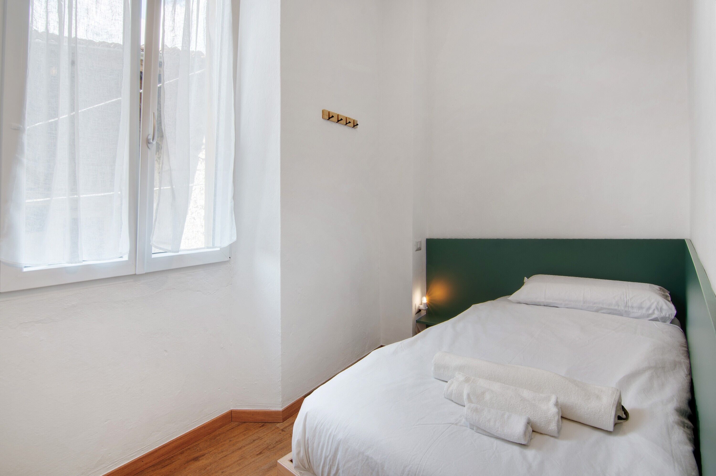 Une chambre à coucher avec un lit, une fenêtre ornée de rideaux légers, une lampe fixée au mur et une petite étagère en bois.