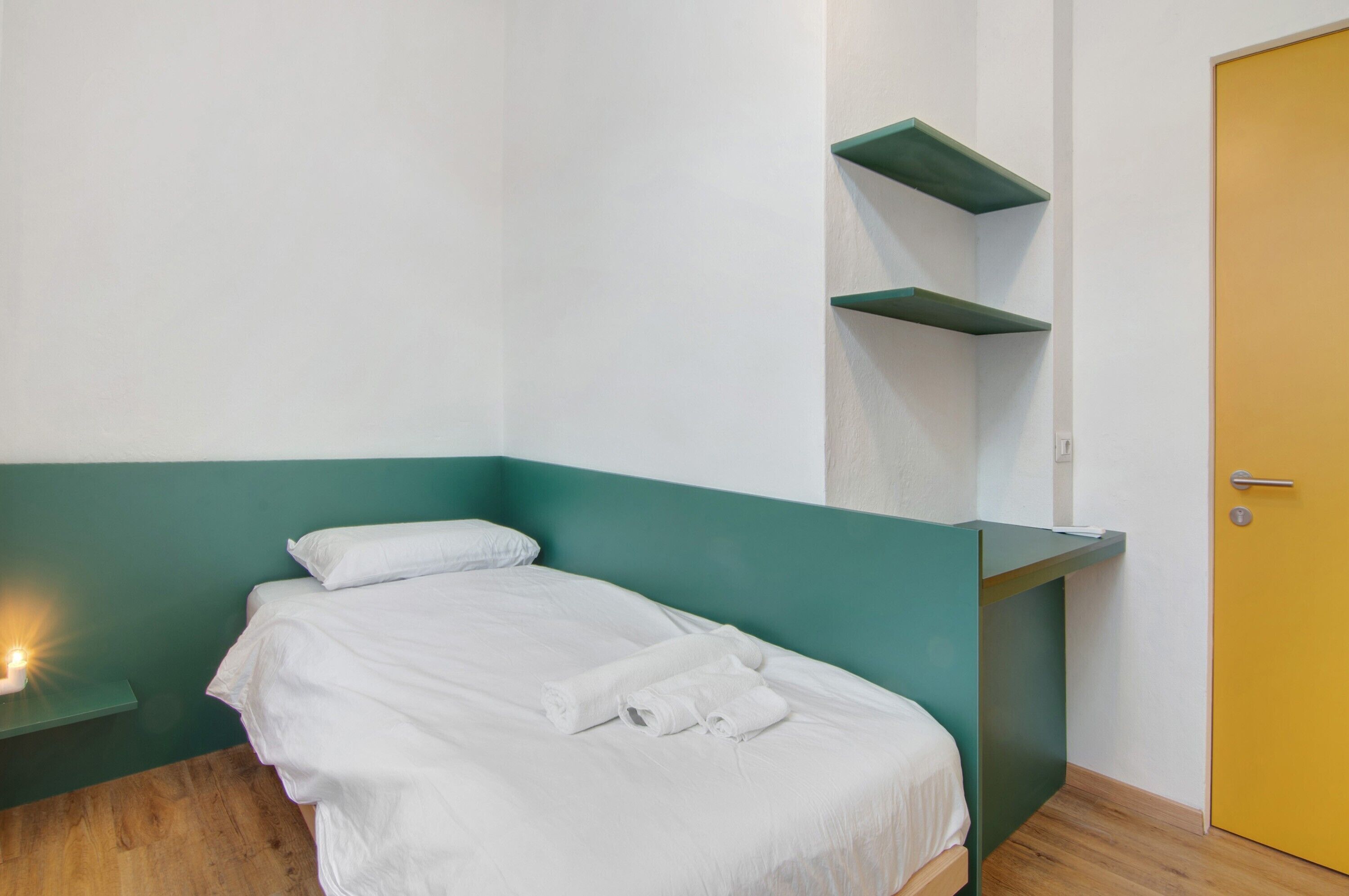 Ein Einzelbett mit weißer Bettwäsche, eine Wand in Grünton, ein Holzboden und eine gelbe Tür.