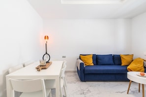 A white dining table with chairs, a blue sofa with yellow cushions, and a small white table with a plate and an orange.