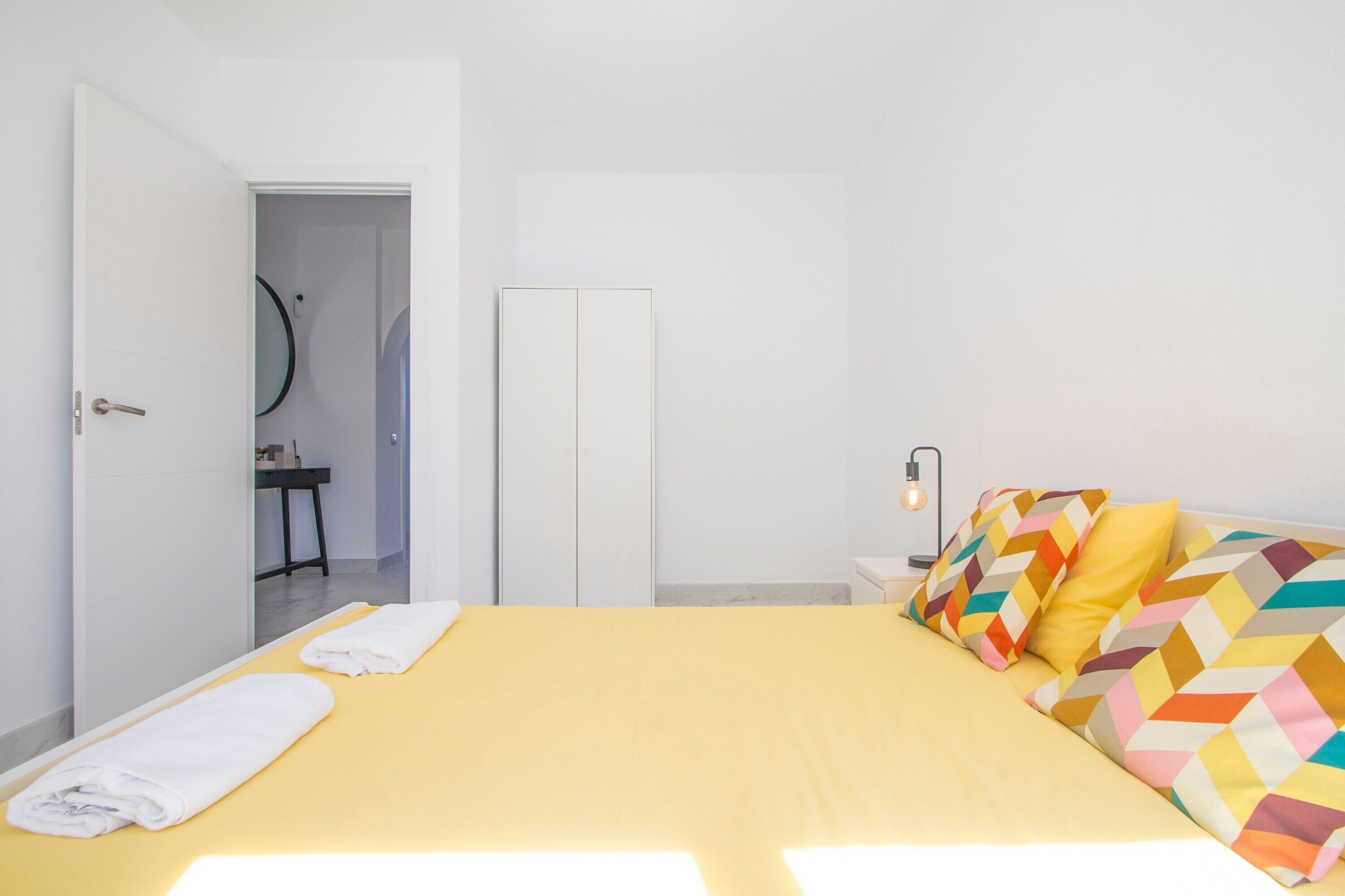A bedroom with a bed, a bedside table with a lamp, a mirror, and a door leading to a bathroom.