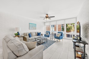 A spacious living room with a white brick wall, a ceiling fan, a large window with a view of greenery, and a blue armchair.