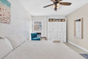 Quarto com cama, cadeira, guarda-roupa, ventoinha de teto e janela.