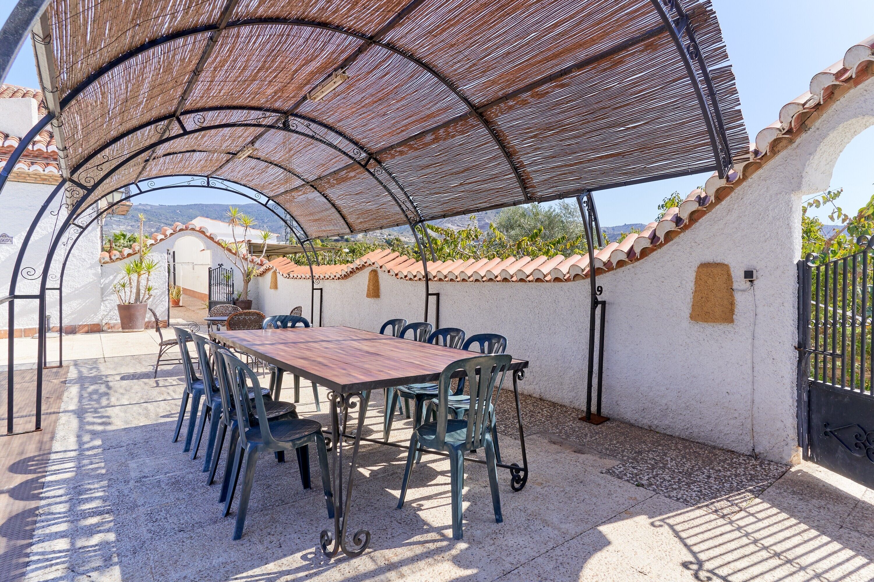 Un área de comedor al aire libre cubierta con mesa y sillas, una pérgola en la parte superior y una pared blanca con tejado de azulejos.