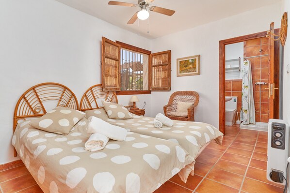 A bedroom with two beds, a ceiling fan, a window with wooden shutters, a wicker chair, and a shower area. - Holiday Home 'Cabrera' with Sea View, Private Pool and Wi-Fi (Puntalon)