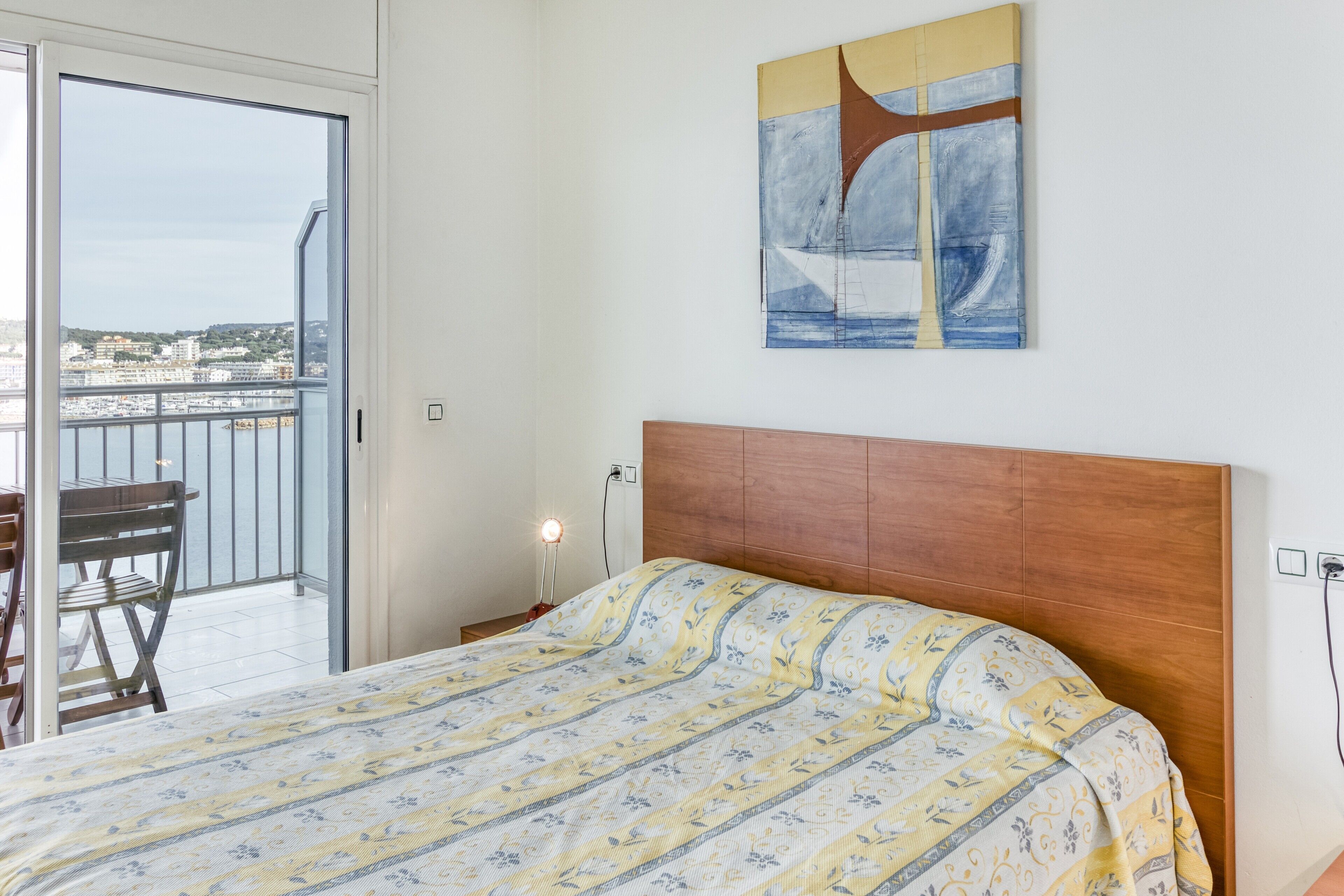 Un dormitorio con cama, cabecera de madera, un cuadro y balcón con vistas a edificios.