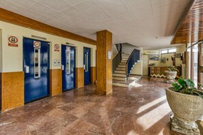 Een hotel lobby met marmeren vloeren, blauwe deuren met de tekst 'ASCENSOR' en een trap.