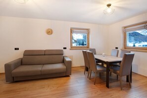 Una habitación con sofá, mesa de comedor, sillas, un reloj y ventanas con vistas nevadas.