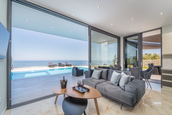 A modern living room with a glass wall overlooking a pool and the sea, a sofa, a table with a tray, and a wine glass.