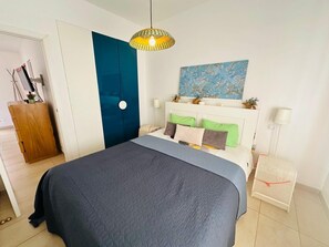 A bedroom with a bed, a blue door, a hanging light fixture, a painting, and a small white table.