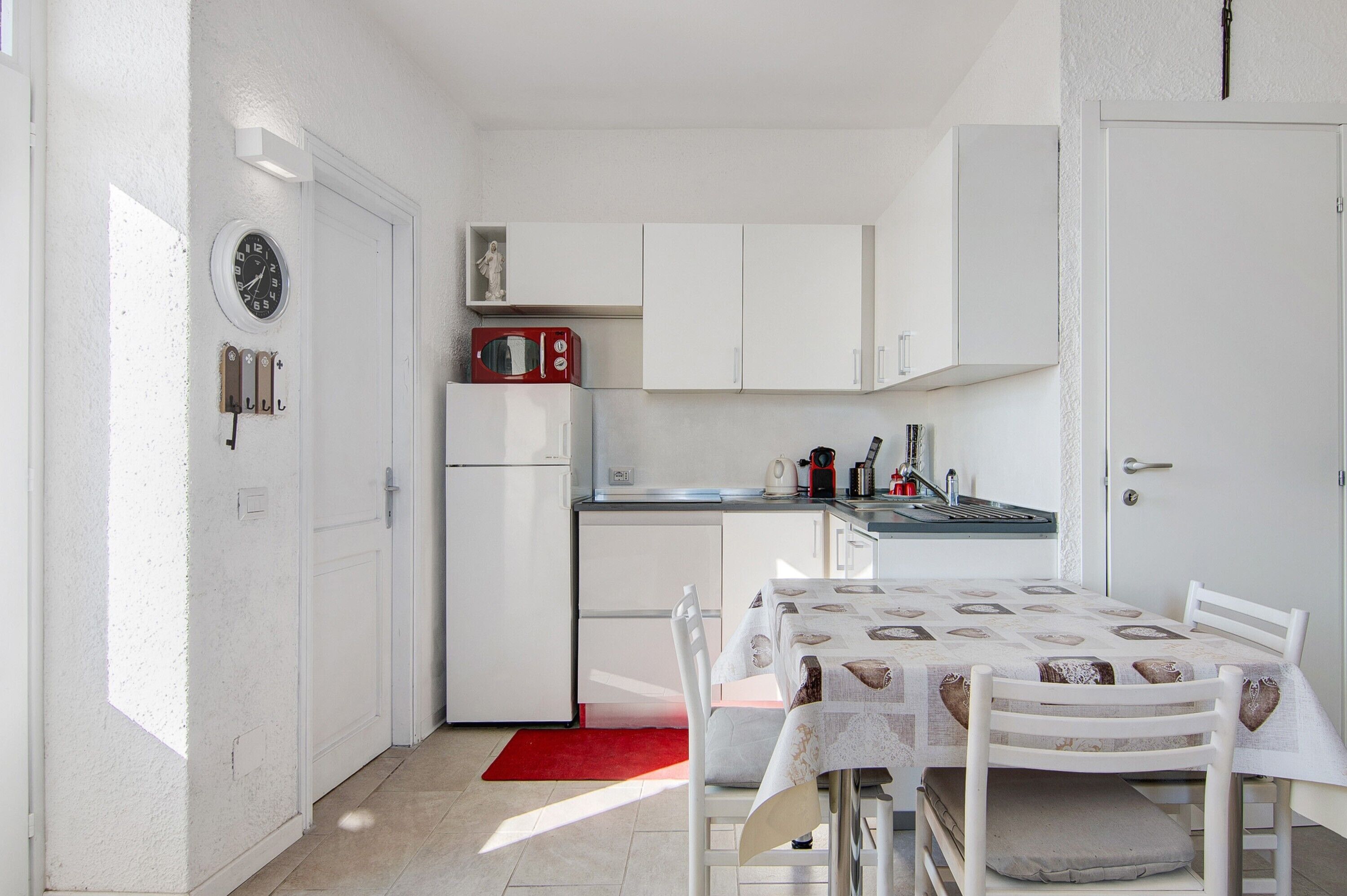Une cuisine de taille réduite avec des meubles blancs, un micro-ondes rouge et une table à manger recouverte d’une nappe à motifs.