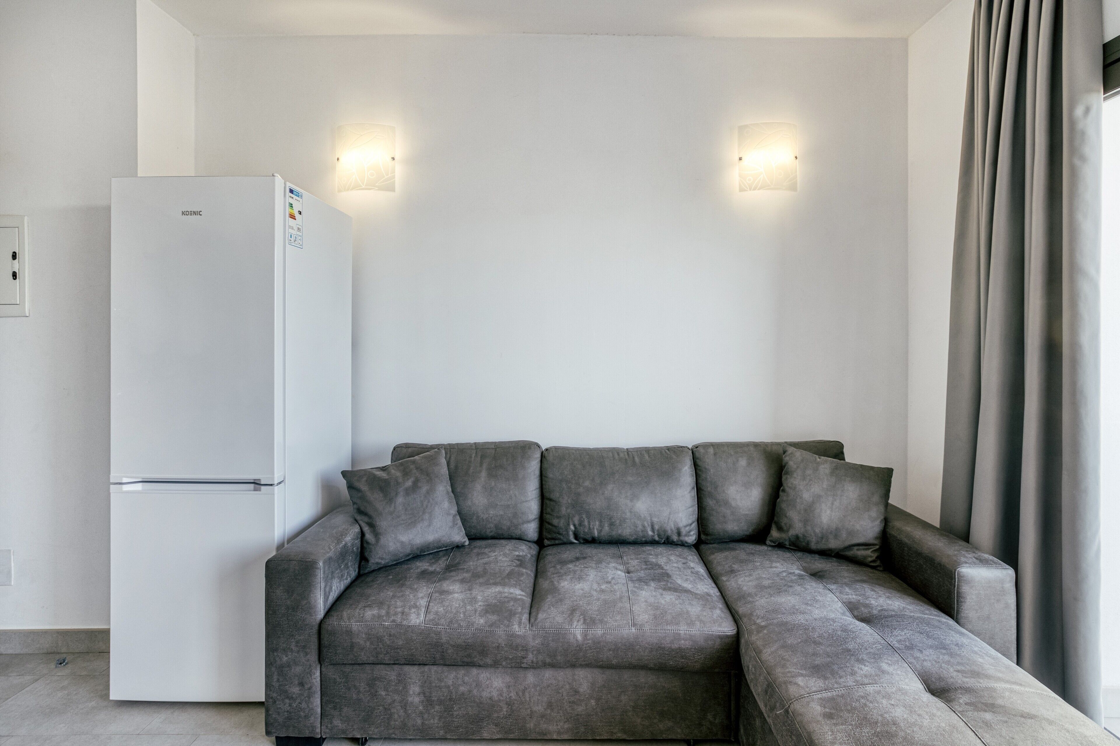 Un salon moderne avec un canapé gris, un réfrigérateur encastré blanc et deux lampes fixées au mur.