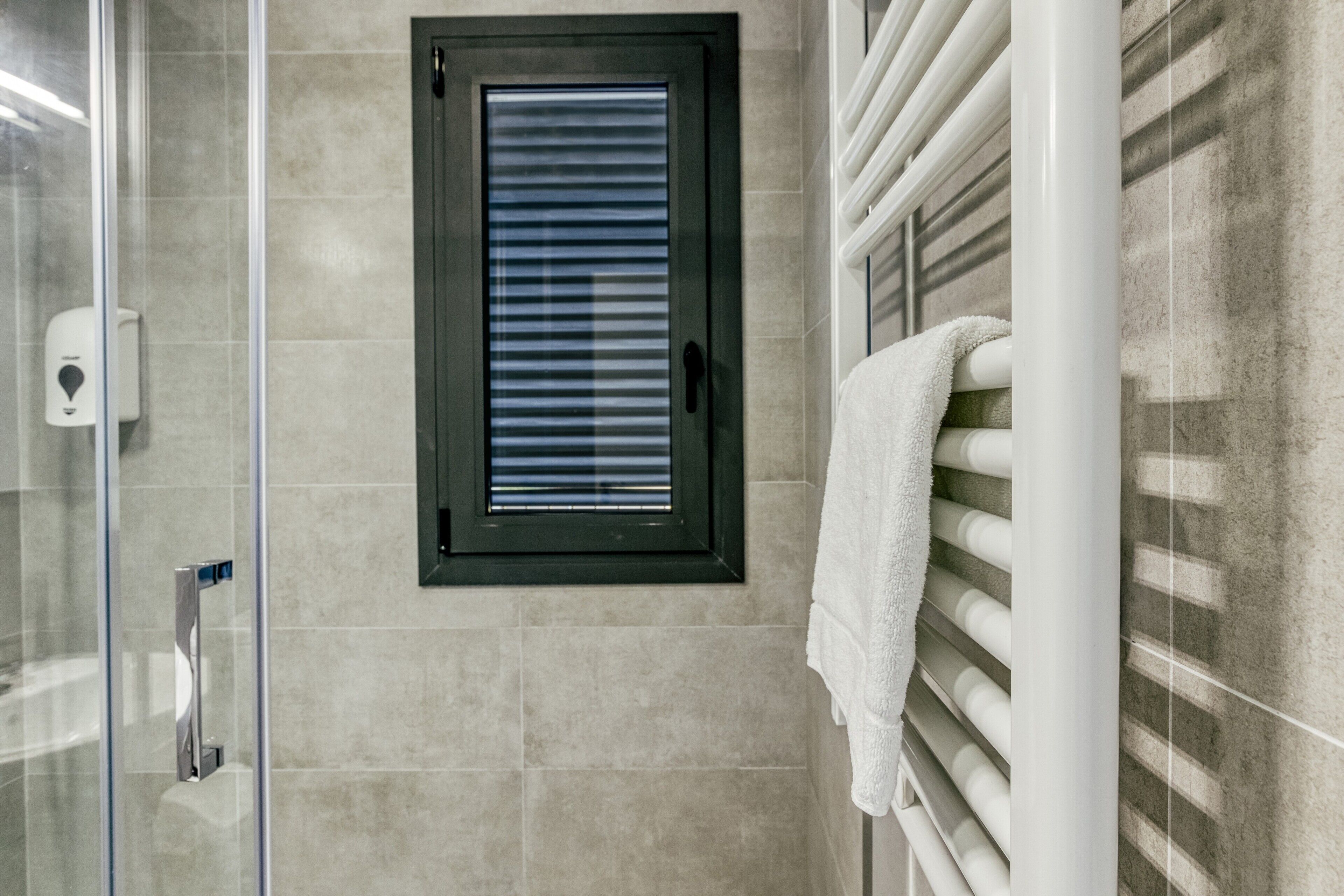 Un baño con cabina de ducha de vidrio, un perchero con una toalla blanca y una ventana con persianas.