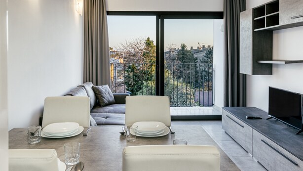 A modern living room with a dining area, a sofa, a TV, and a balcony with a view of buildings and trees.