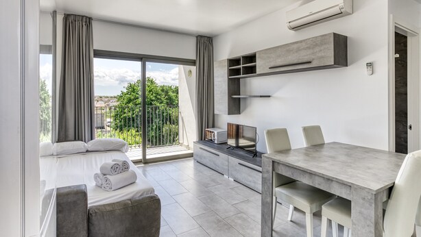 A modern room with a bed, a dining table, chairs, a TV, and a balcony with a view.