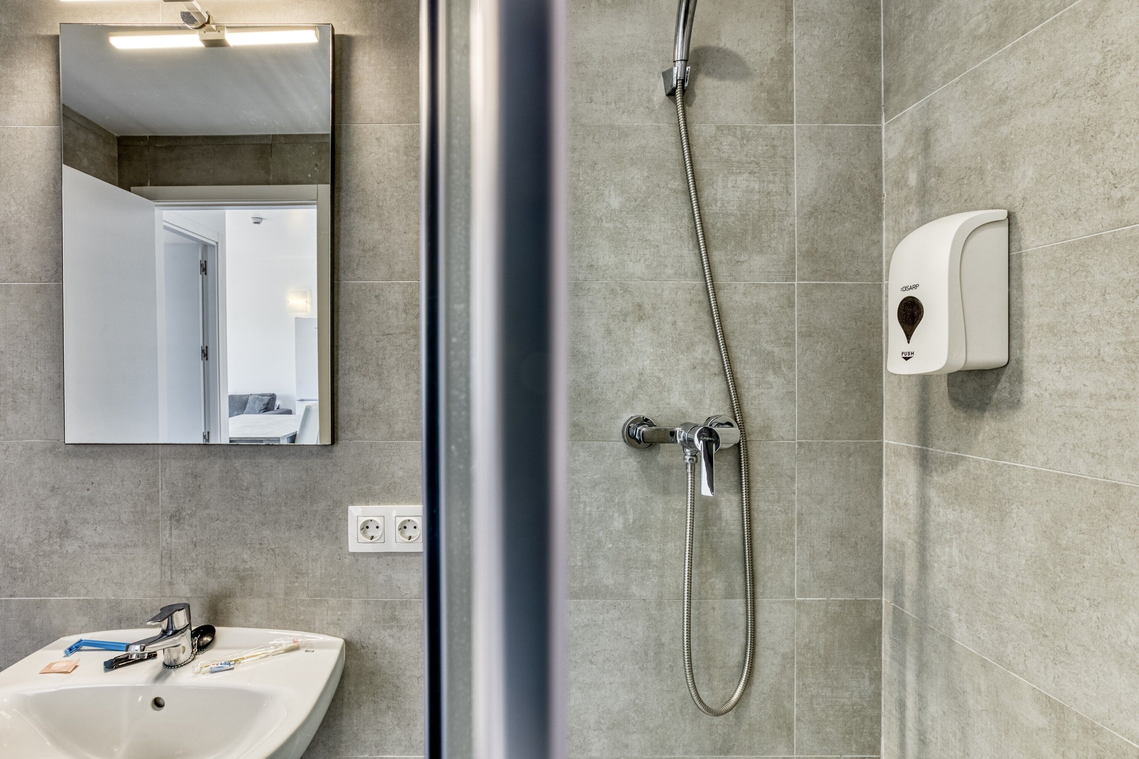 A bathroom with a sink, mirror, shower, and soap dispenser.