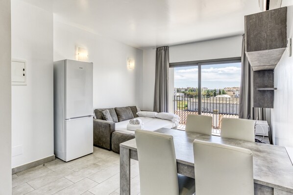 A modern kitchen with a built-in refrigerator, a dining area with a table and chairs, a sofa, and a large window with a view of the city.