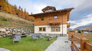 A two-story wooden chalet with a balcony, outdoor seating, and a stone wall. - Apartment 'Palipert Flowers House' with Mountain View, Shared Garden and Wi-Fi (Livigno)