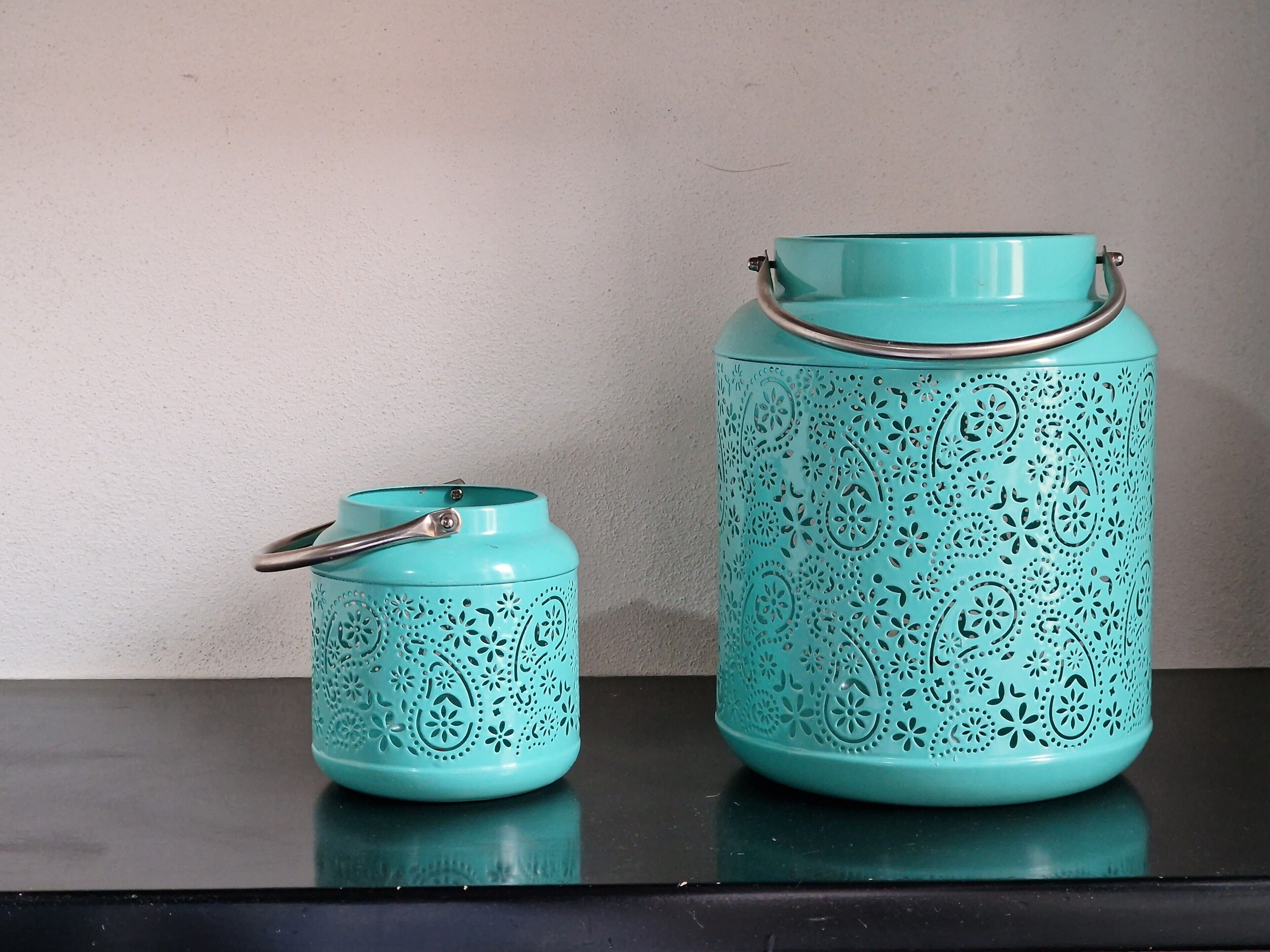 Dos recipientes azul turquesa con diseños intrincados en relieve y asas metálicas, sobre una superficie reflectante frente a una pared lisa.