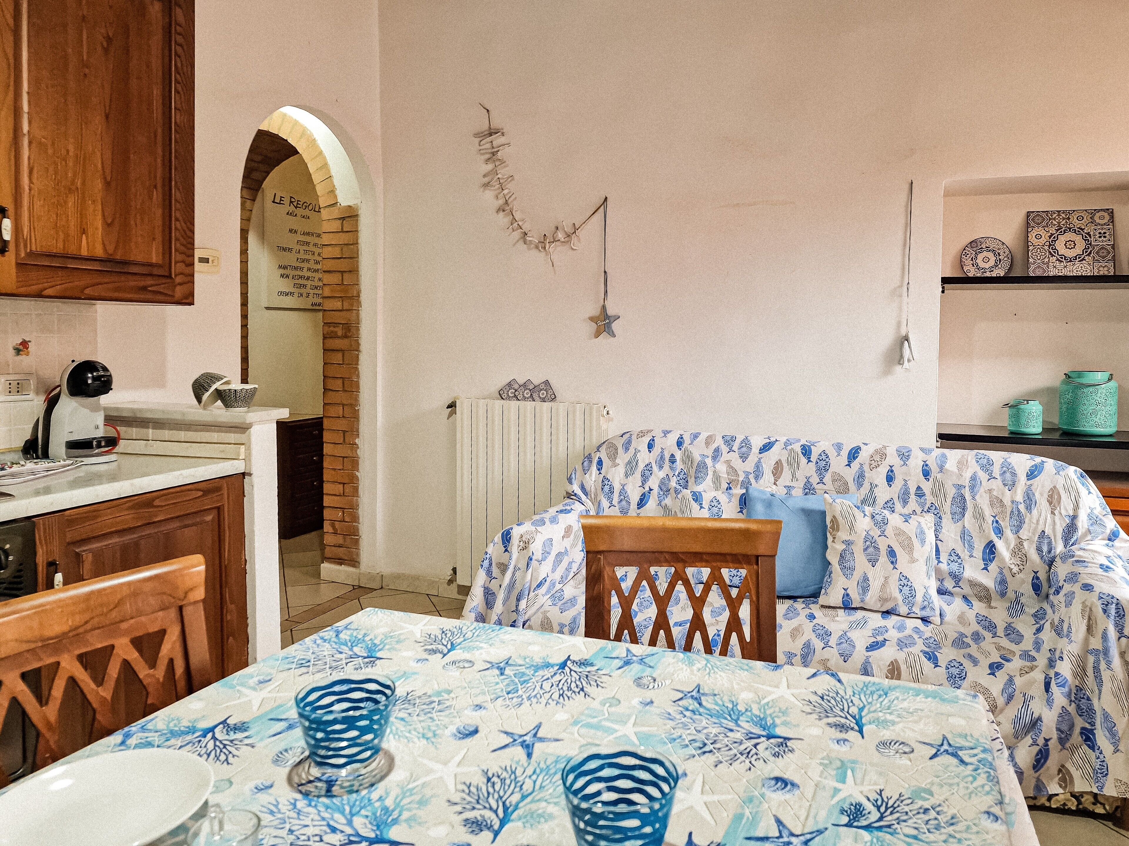 Ein Zimmer mit einem Bett, das mit einer blau-weißen Tagesdecke bezogen ist, einem Holzkopfstück, einem Tisch mit einer blau-weißen Tischdecke, einer Küchenzeile mit Holzausstattung und einer Backsteinwand mit einem Bogen.