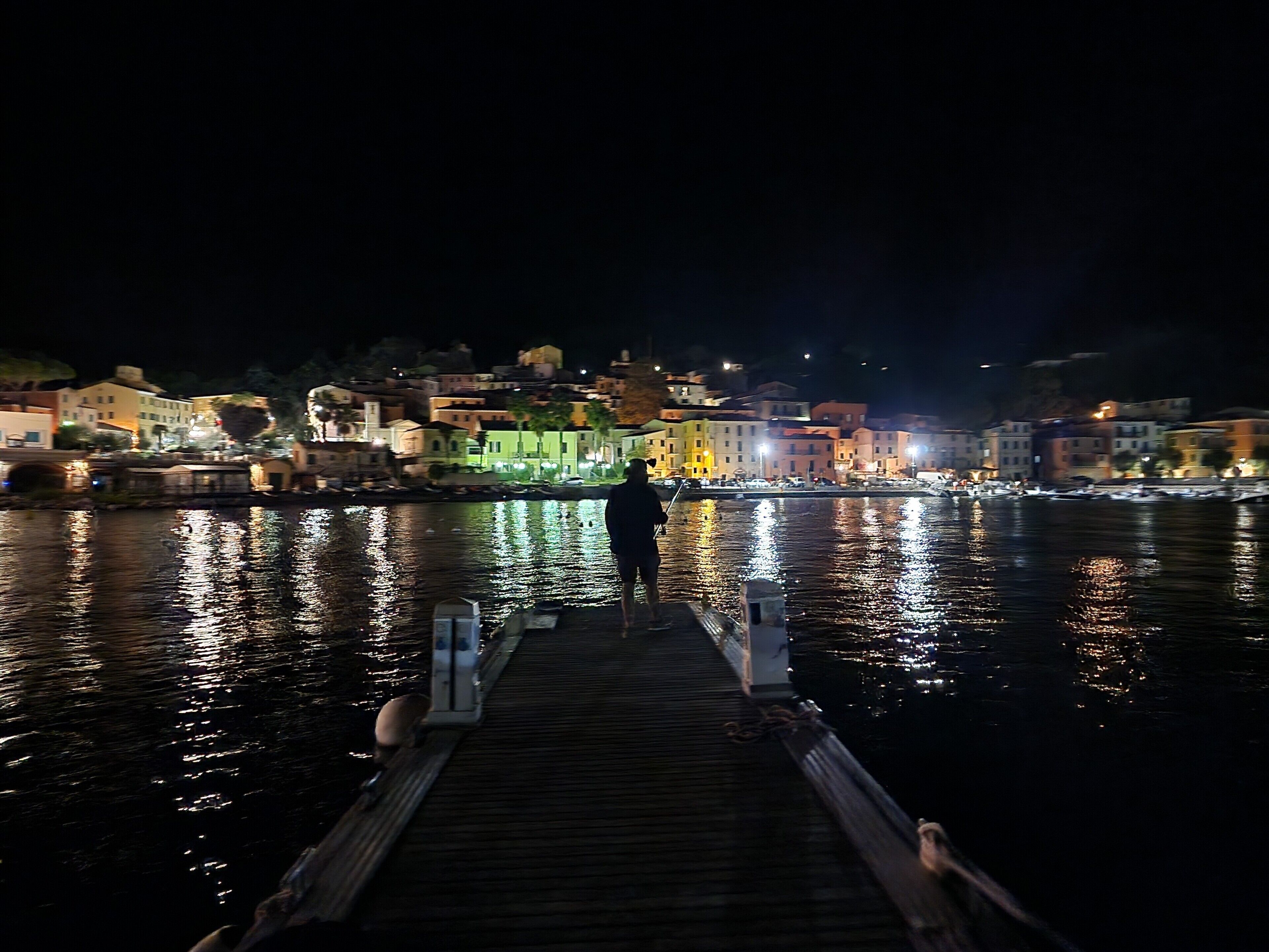 Une scène nocturne au bord de l’eau, avec une jetée s’étendant dans une eau calme, des bâtiments dont les lumières se reflètent sur l’eau, et une personne debout au bout de la jetée.