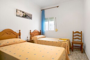 Ein Zimmer mit zwei Betten, einem Holzsessel, einem Nachttisch, einer Lampe, einem Gemälde und einem Fenster mit Vorhängen.