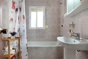 Ein Badezimmer mit einer Badewanne, einem weißen Waschbecken, einem Holzboden mit Handtüchern und einem Fenster mit weißem Rollo.