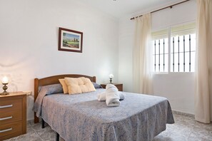 Ein Schlafzimmer mit einem Bett, einem Nachttisch, einer Lampe, einem Fenster mit Vorhängen und einem Bild an der Wand.