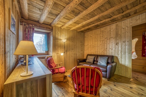 A wooden cabin interior with a wooden desk, a red chair, a leather sofa, and a window with curtains. - Apartment 'Anna-Purna' with Mountain View, Shared Terrace and Wi-Fi (Arâches-la-frasse)