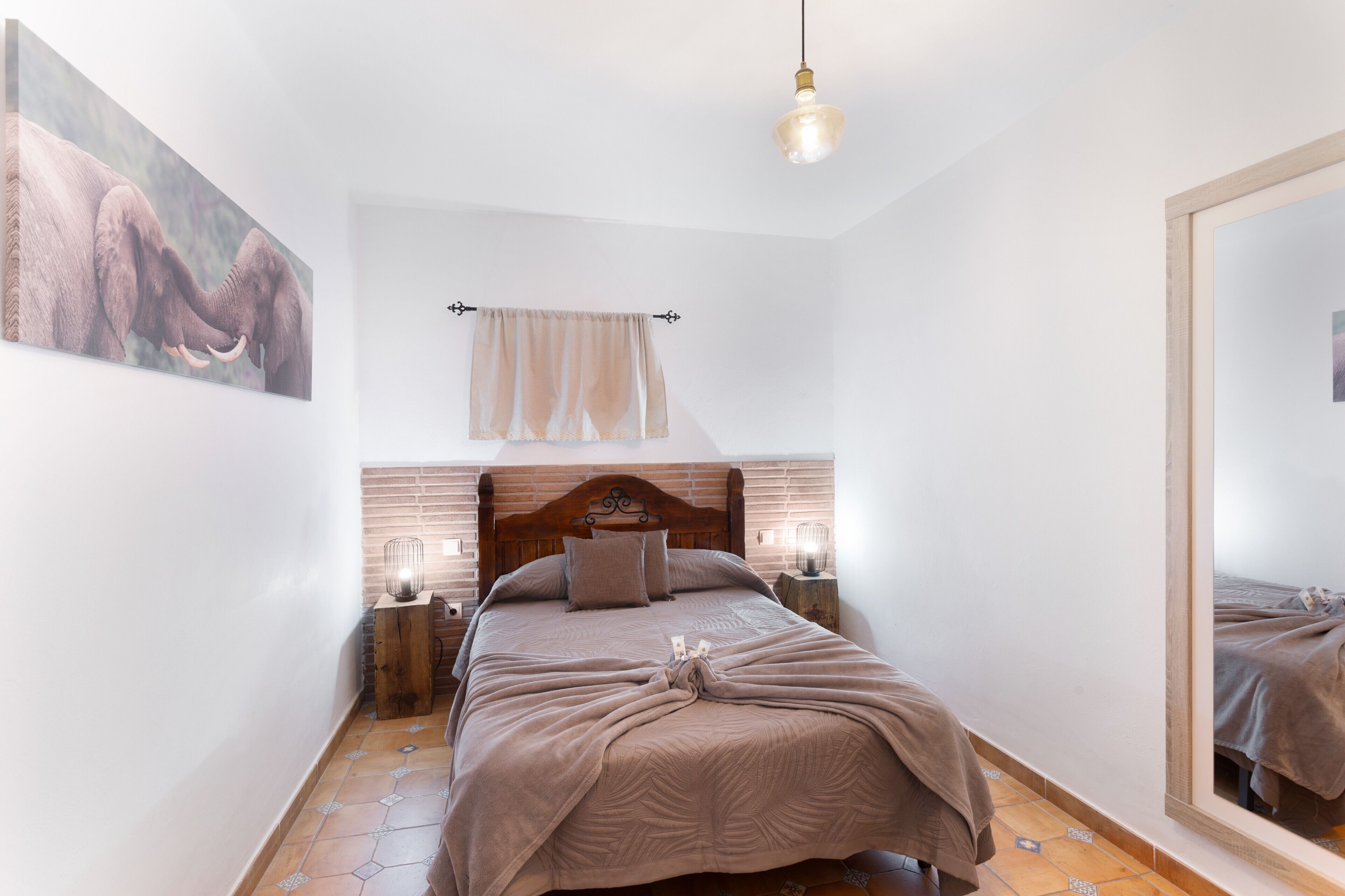 Een slaapkamer met een houten hoofdbord, een bed met een beige deken, twee bedlampjes, een hanglamp, een grote spiegel en een schilderij aan de muur.