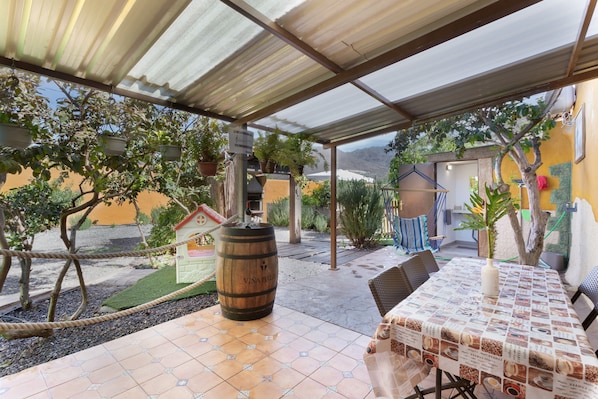 A covered patio area with a table set for dining, surrounded by potted plants and a wooden barrel.
