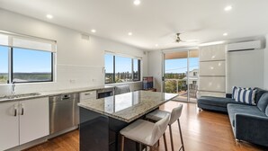 A modern kitchen with a large island, built-in appliances, and large windows offering a view of the landscape. - Mingara 4 - Panoramic River, Ocean & Sunset Views - Holiday Yamba (Yamba)