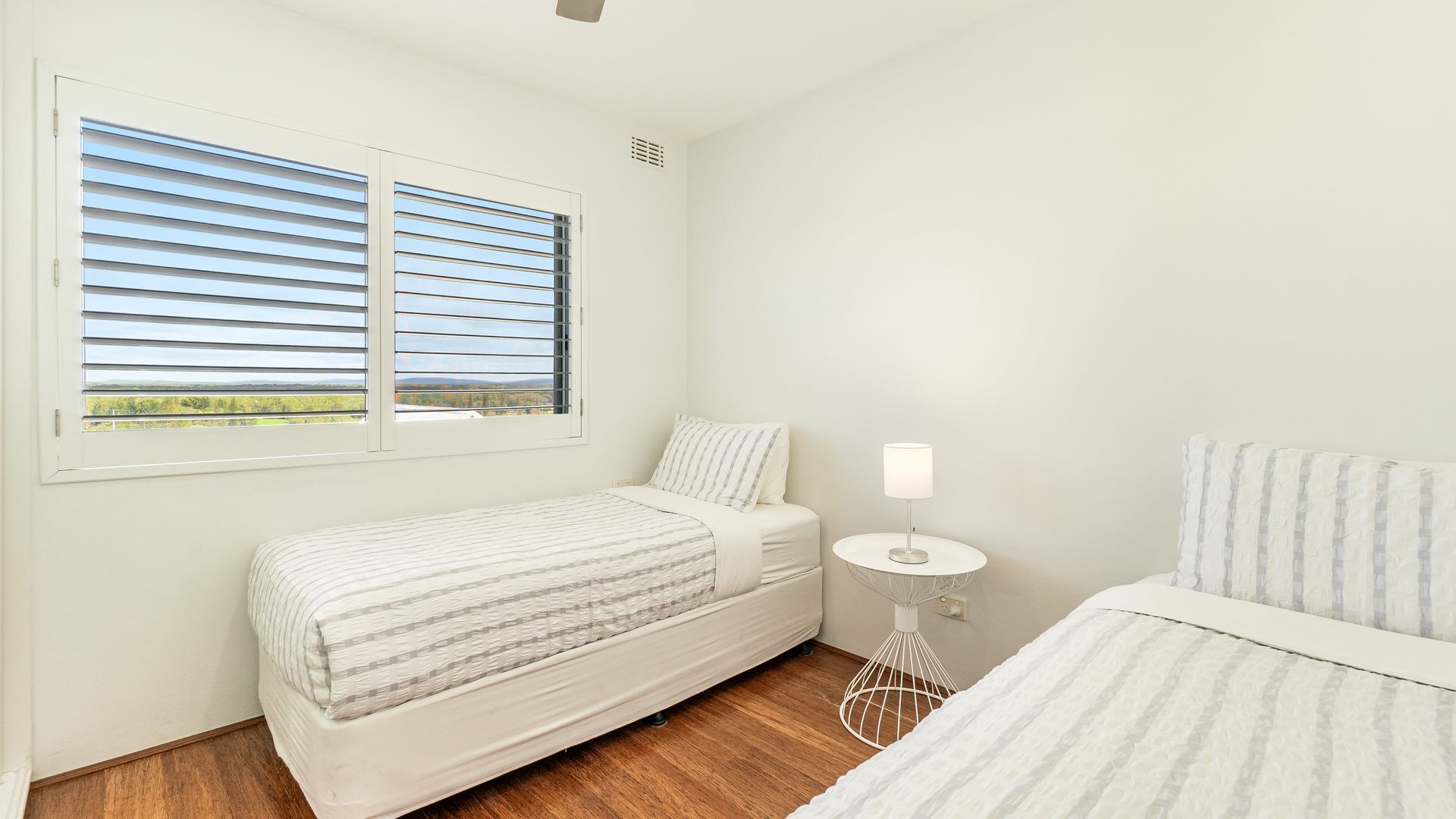 Un dormitorio con dos camas, una mesita de noche con lámpara y una ventana con persianas horizontales.