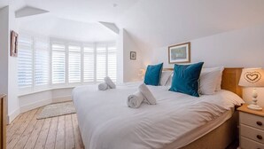 A bedroom with a large bed, white bedding, a nightstand with a lamp, and a window with blinds. - The White House, Southwold (Southwold)
