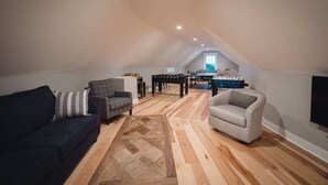 Un espace de vie de style loft avec un plafond haut, un plancher en bois, un canapé foncé, un fauteuil gris et une table à manger agrémentée d’un baby-foot.