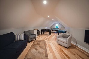A loft-style living space with a high ceiling, wooden flooring, a dark sofa, a grey armchair, and a dining table with a foosball table. - Ann Riley Farmhouse - Country Setting By The Horse Show (Williamsburg)