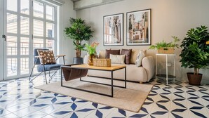 A living room with a sofa, armchairs, a coffee table, and potted plants. There are two framed pictures on the wall.