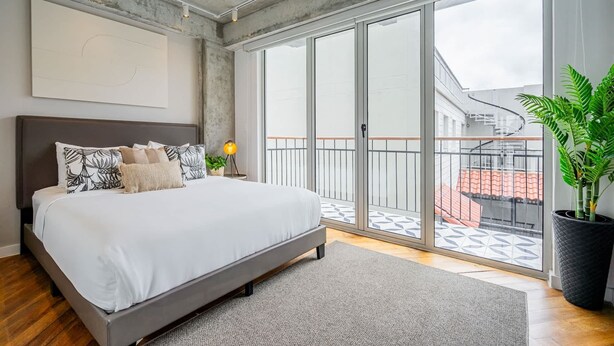 A modern bedroom with a large bed, a view of the city through floor-to-ceiling windows, and a balcony with a railing.
