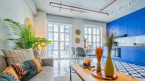 A modern living room with a sofa, coffee table, and potted plants. There is a dining area with chairs and a table. The kitchen has blue cabinets and a stove. - w* | Extraordinary 2BR in Plaza Herrera (Panamá)