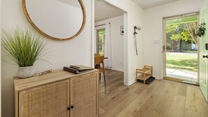 Un corridoio con pavimento in legno, uno specchio rotondo, una pianta in vaso, una piccola sedia di legno e una porta che dà all'esterno.