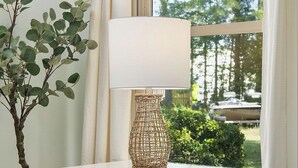 Una camera da letto con comodino, lampada, una piccola pianta e una finestra che si affaccia su alberi e un recinto.