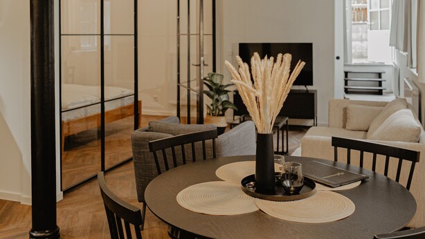 A dining area with a round table, black chairs, a black pendant light, and a vase with dried plants.