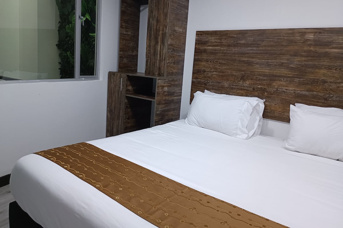 A neatly made bed with white bedding and a brown patterned throw, a wooden headboard, and built-in shelving with a green plant.