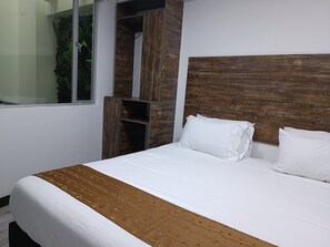 A neatly made bed with white bedding and a brown patterned throw, a wooden headboard, and built-in shelving with a green plant.