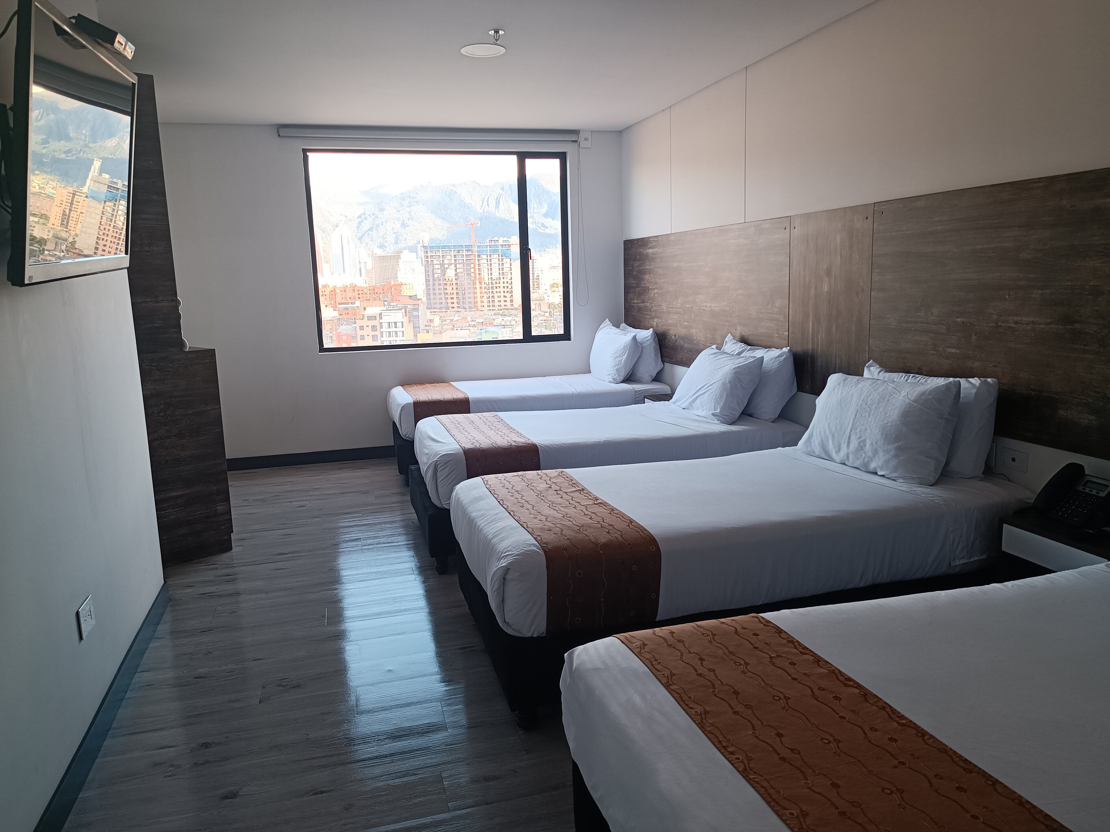 Habitación de hotel con tres camas, un ventanal y vistas a la ciudad, y una pared decorativa de madera.