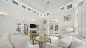 A spacious living room with a high ceiling, white walls, and large windows. There is a wooden coffee table, white sofas, and a flat-screen TV mounted on the wall. - Designer Home in SeasideFL, 272 feet to beach, + 2 Bikes! (Santa Rosa Beach Fl)
