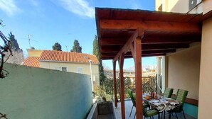 Un espacio de comedor al aire libre cubierto, con una mesa y sillas, vista panorámica de la ciudad y un edificio con tejado de tejas rojas.