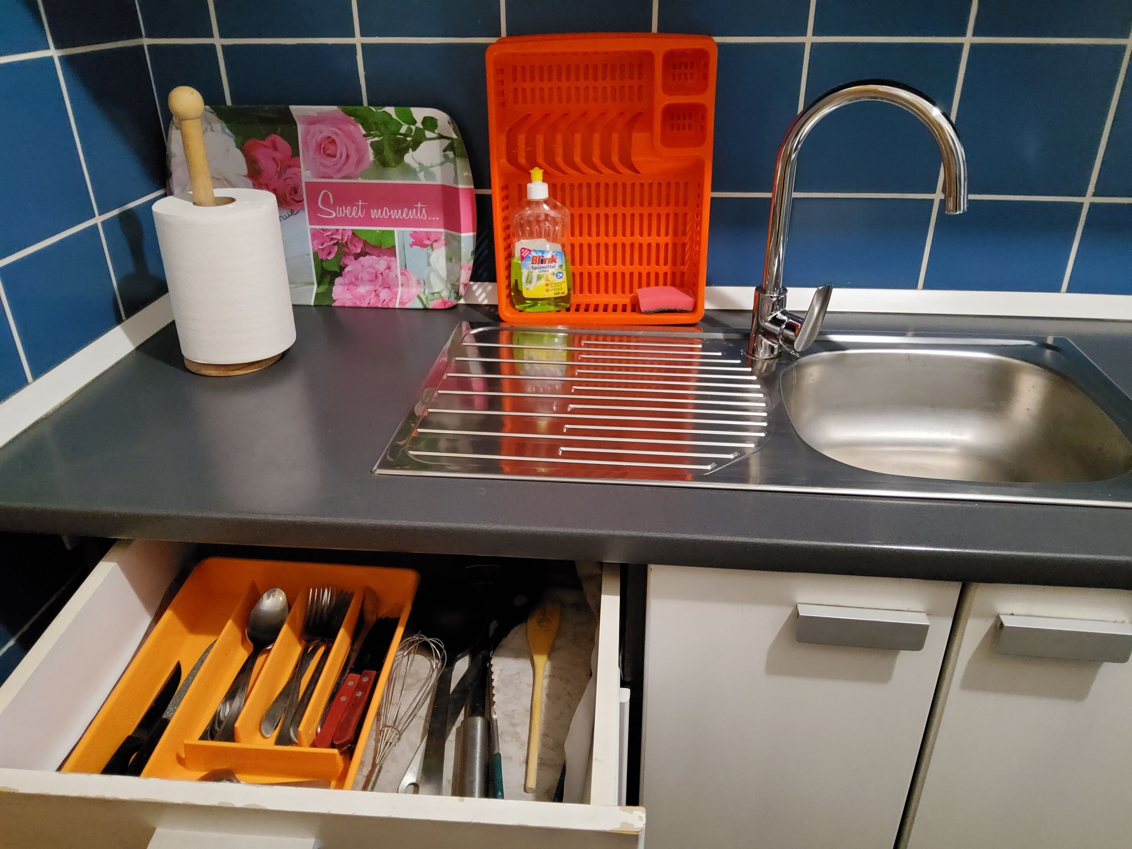 A kitchen with a stainless steel sink, a dish rack, a dish soap bottle, and a set of utensils in a drawer.